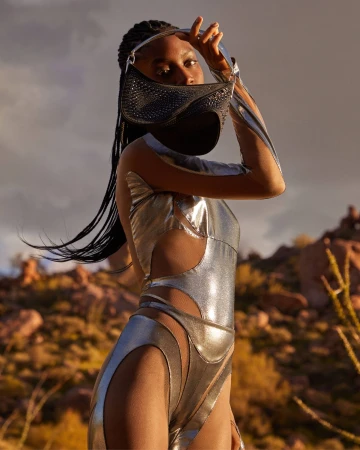 A young adult woman poses in a desert landscape, wearing a reflective silver bodysuit, and holding a sparkly black handbag over her face.