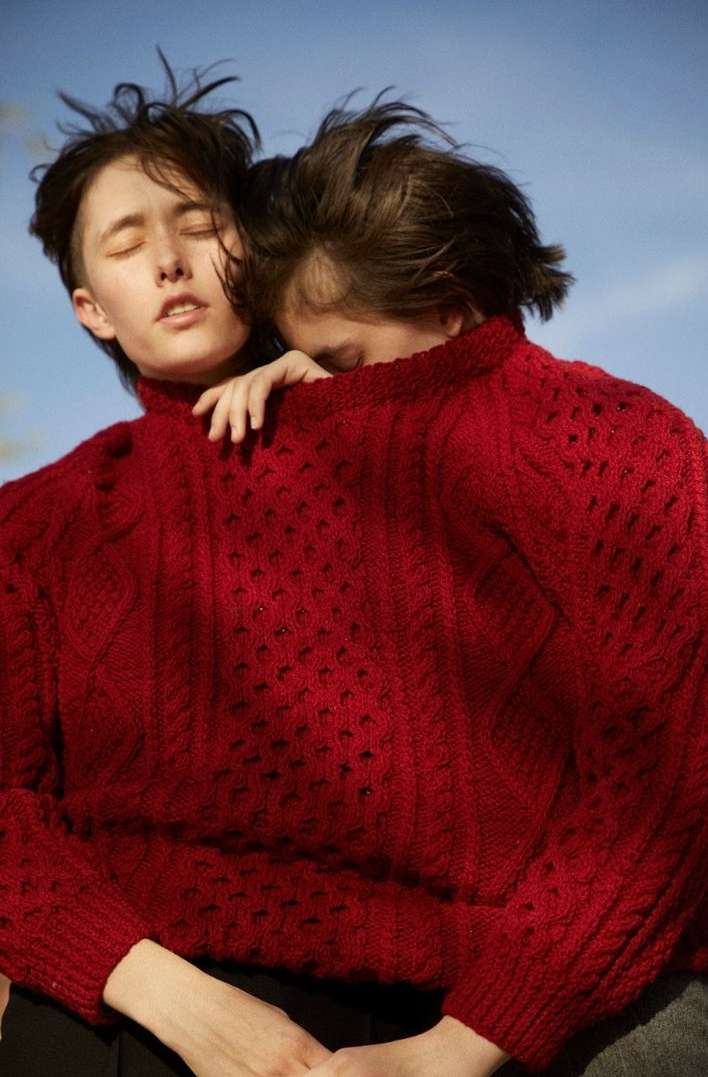 Two people in red sweaters embrace outdoors against a clear sky, one with eyes closed and the other with head resting.