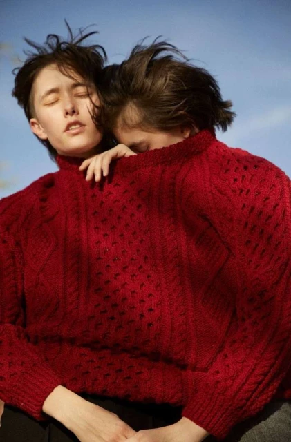 Two people in red sweaters embrace outdoors against a clear sky, one with eyes closed and the other with head resting.