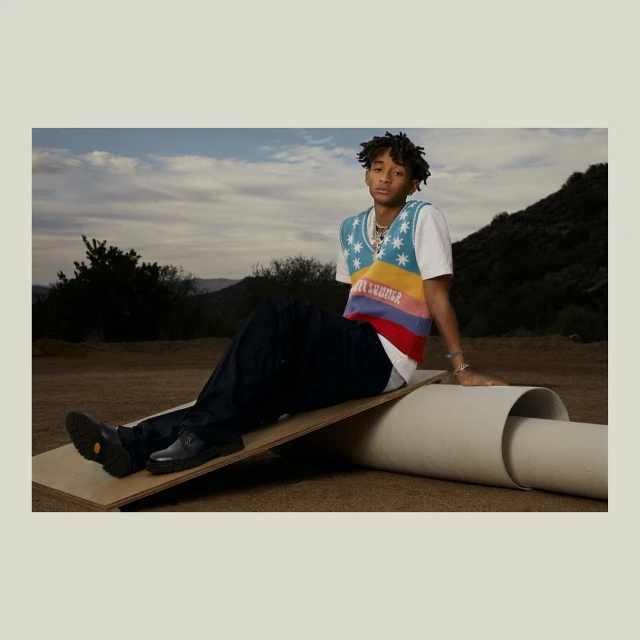 Jaden Smith poses outdoors sitting on a board, wearing a colorful vest, dark trousers, and black shoes. The backdrop features a cloudy sky and mountains.