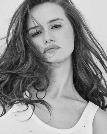 Close-up black and white portrait of a young woman with long flowing hair wearing a white tank top against a plain background
