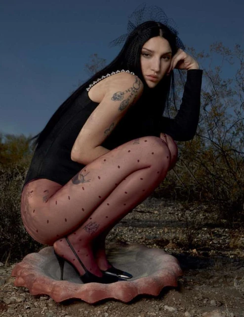 Woman in black bodysuit and polka dot tights squats on concrete basin in desert landscape under blue sky. Beauty fashion shot outdoors.