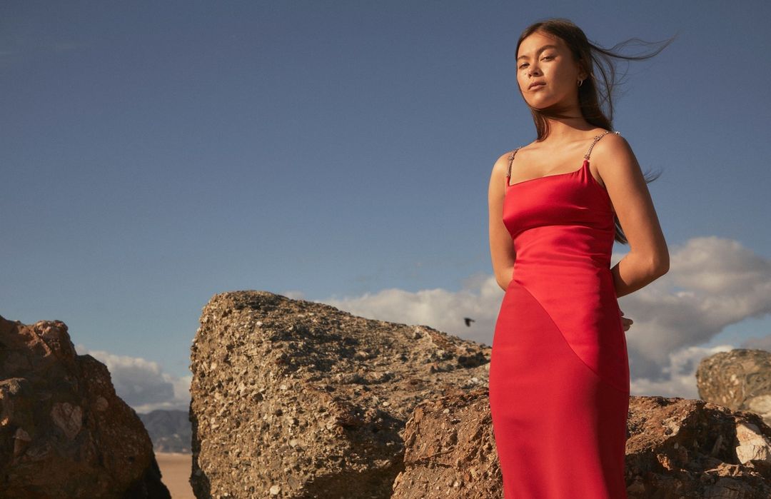 Woman in a red dress standing on rocks against a blue sky backdrop. Fashion shot with natural light on a rocky beach setting.