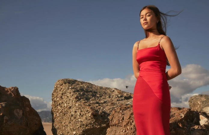 Woman in a red dress standing on rocks against a blue sky backdrop. Fashion shot with natural light on a rocky beach setting.