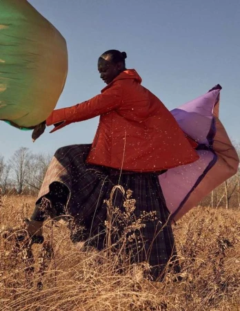 Outdoor fashion shoot featuring a Black woman in a red jacket, plaid skirt, and green and lavender fabric, standing in a golden field.