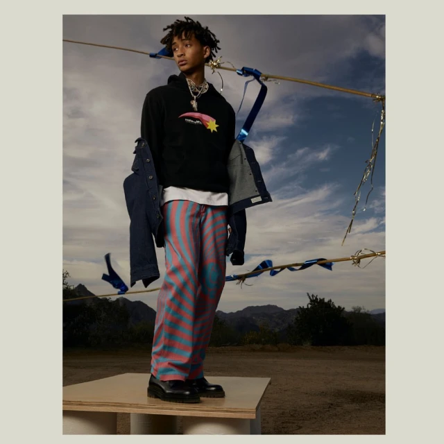 Adult male poses on a platform wearing a black hoodie with a shooting star, vertically striped pants, and black shoes outdoors.