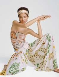 Model wearing a patterned headscarf and matching printed silk top and wide-leg trousers against a plain white studio backdrop.
