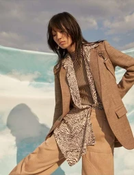 Model with dark hair wearing a beige blazer, patterned blouse, and matching trousers against a cloudy sky backdrop.