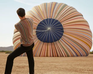 Man pulling a rope attached to a giant striped hot air balloon resting on the ground in a desert landscape