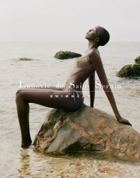Model wearing olive green bikini seated on a wet rock in shallow water with rocky coast background
