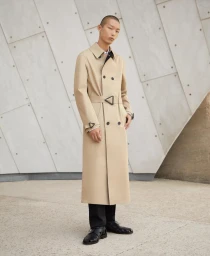 Model wearing a long beige trench coat with black trousers and black shoes outdoors against a geometric concrete panel wall.