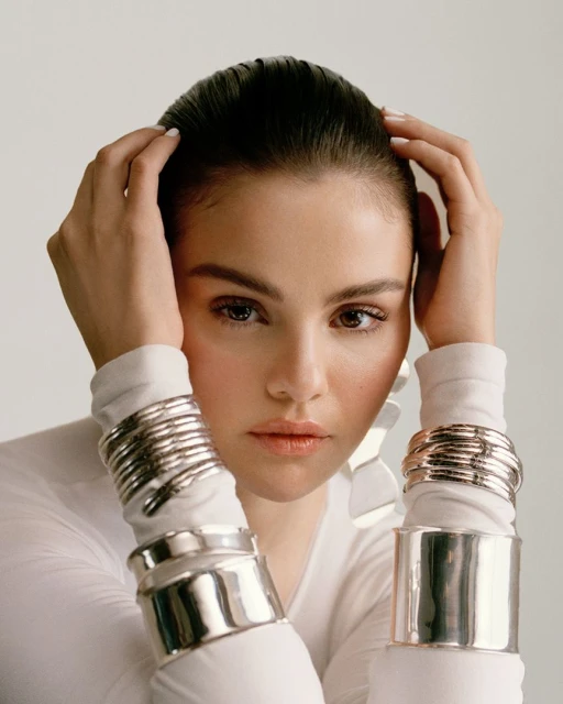 Close-up of an adult female model wearing shiny silver bangle bracelets on both arms, hands near her head, in a photo studio.