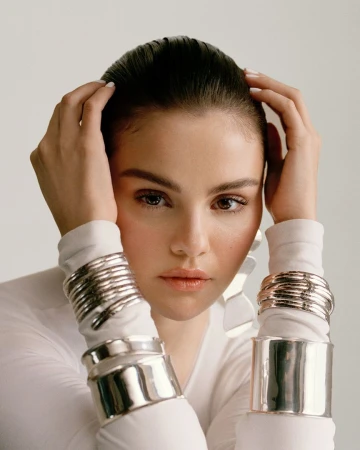 Close-up of an adult female model wearing shiny silver bangle bracelets on both arms, hands near her head, in a photo studio.
