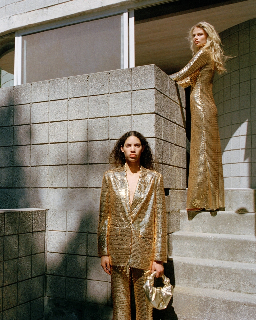 Two women pose outdoors wearing matching shimmering gold sequin pant suits and dress against a grey concrete block building background.