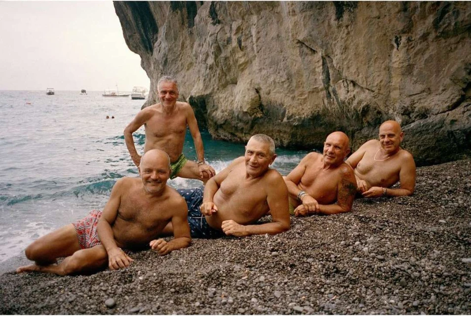 A group of five shirtless mature men pose on a rocky beach, with ocean water and a large rock formation in the background during daylight.