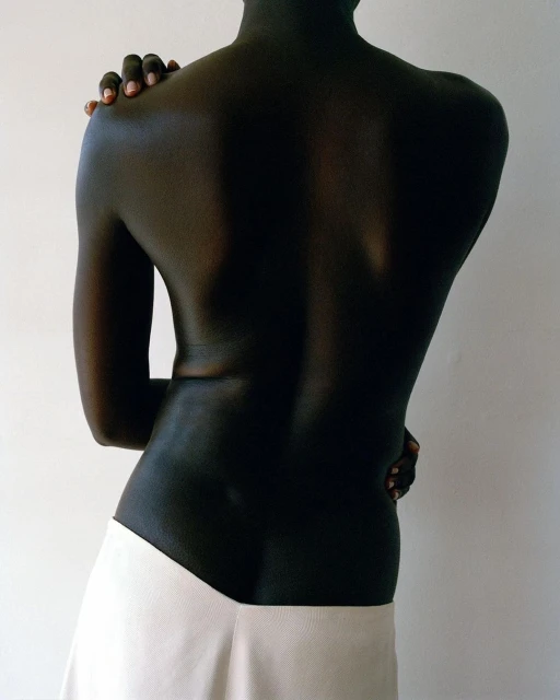 Close-up studio shot of a slender, dark-skinned person with arms crossed, wearing a white skirt against a plain, light-colored background.