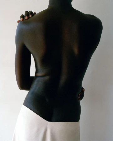 Close-up studio shot of a slender, dark-skinned person with arms crossed, wearing a white skirt against a plain, light-colored background.