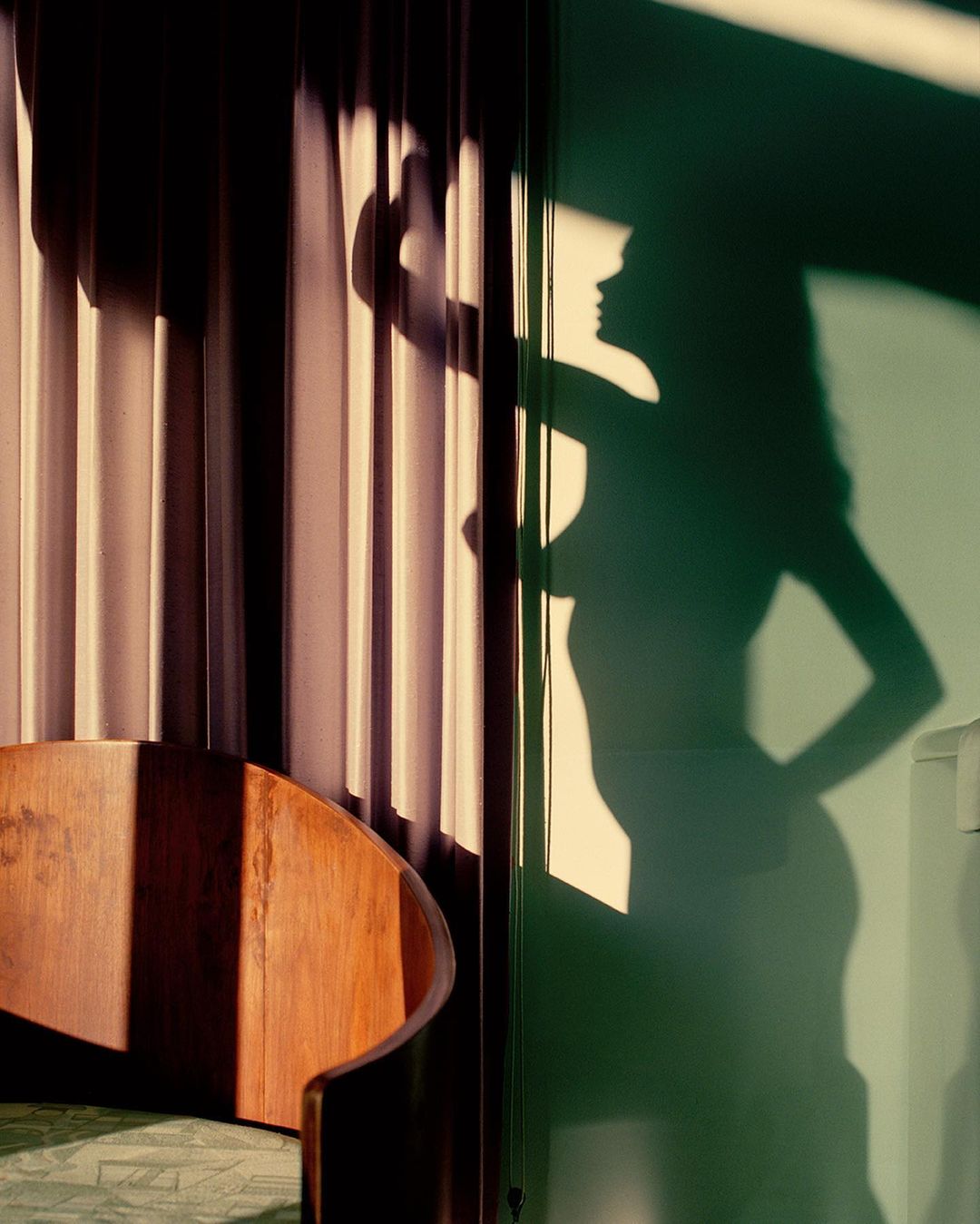Moody interior shot with a silhouetted woman and soft light, featuring a wooden chair, window light, and draped curtains.