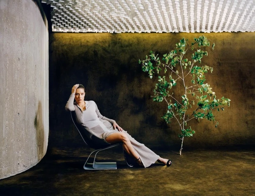 Adult woman poses in a grey dress on a modern chair near a wall, complemented by a small tree against a dark backdrop.