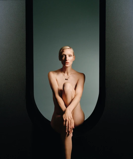 Nude woman with short blonde hair sits in front of a grey background framed by a matte black archway.