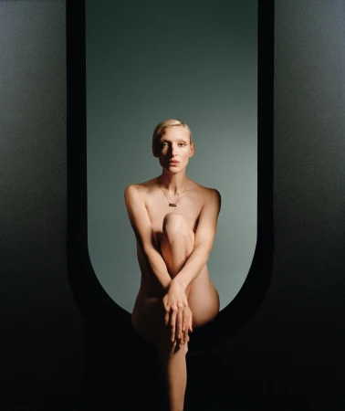 Nude woman with short blonde hair sits in front of a grey background framed by a matte black archway.