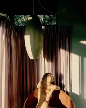 Woman with long blonde hair seen from behind, leaning over a wooden curved chair in a room with draped curtains and a large pendant light.