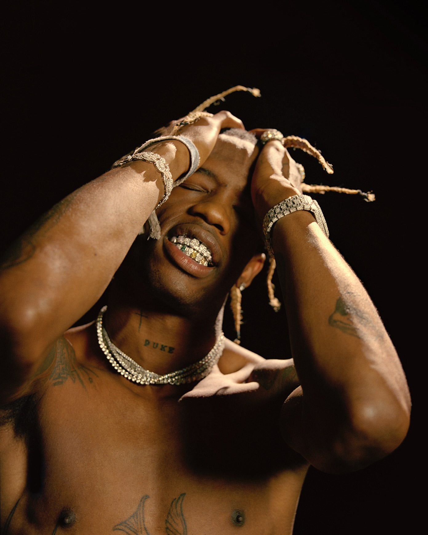 Close-up studio portrait of a shirtless man with light skin, jewelry, grill, and dreadlocks, posing against a dark background.