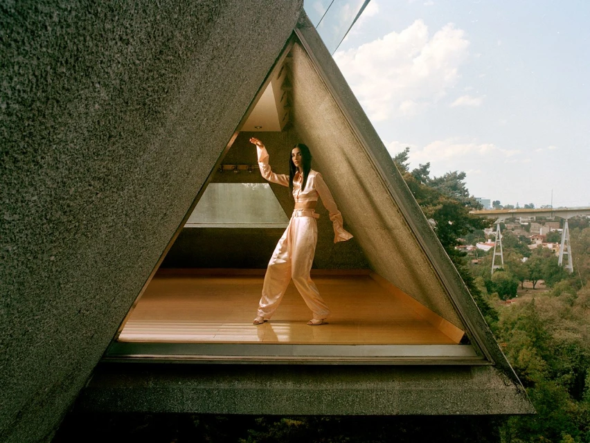 Fashionable woman in loose pink suit strikes dynamic pose in a triangle doorway with a bridge and trees in the background.
