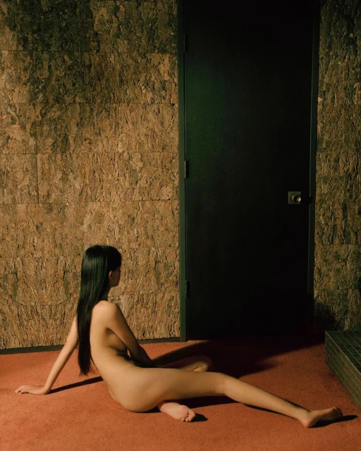 An adult female sits nude on a red carpet indoors against a textured wall and black door in a dimly lit room, looking away.