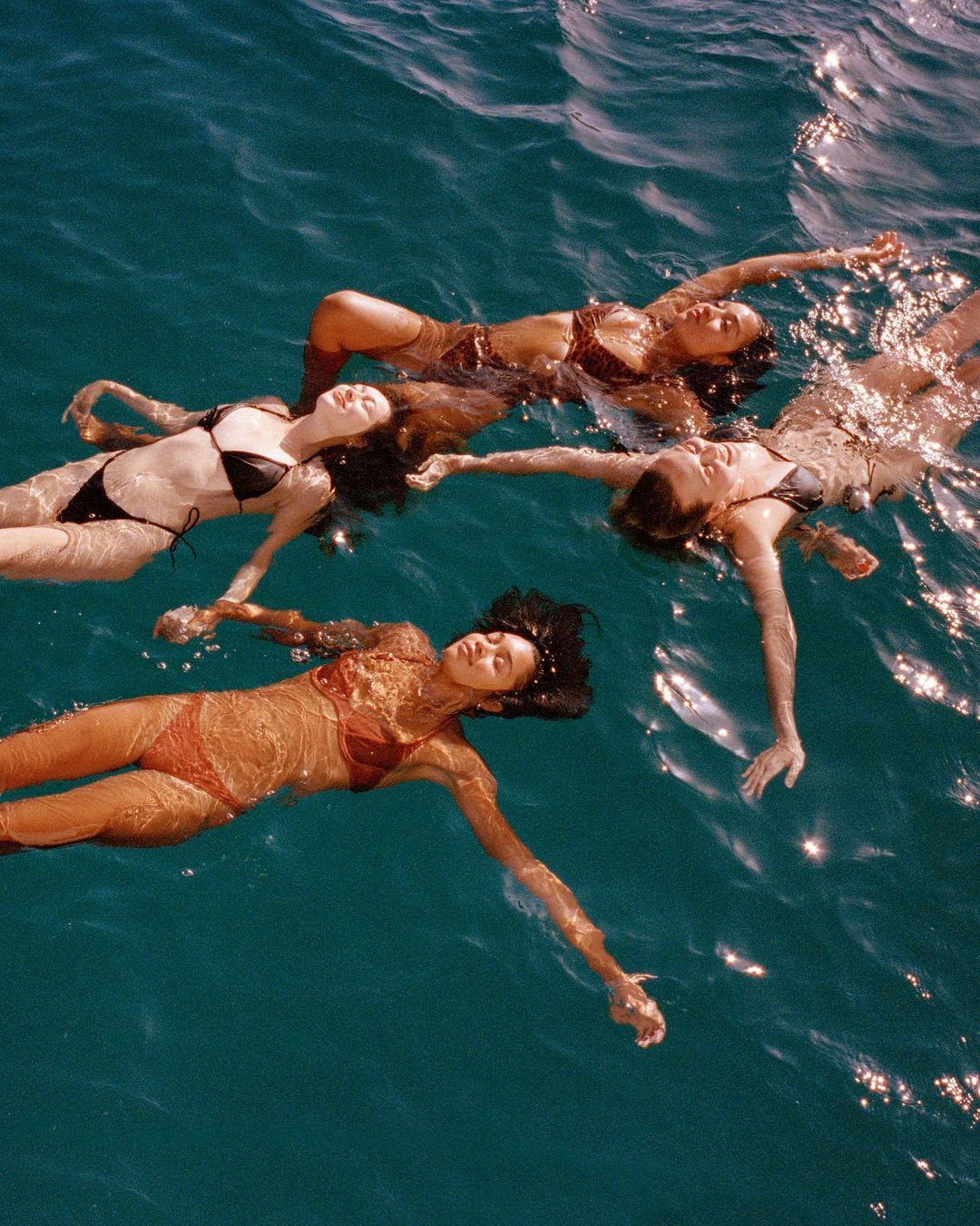 Four women float on their backs in the ocean, wearing swimsuits in various colors. Sunlight reflects on the water around them.