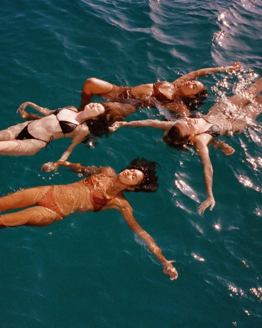 Four women float on their backs in the ocean, wearing swimsuits in various colors. Sunlight reflects on the water around them.
