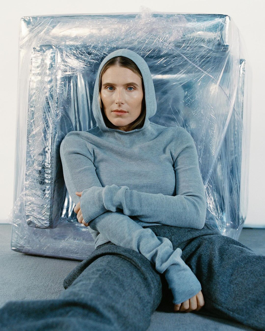 Adult woman in grey hooded knit sweater and sweatpants sitting indoors with silver wrapped object in background. Studio lighting.