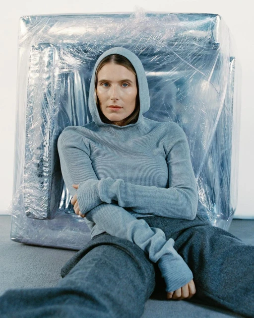 Adult woman in grey hooded knit sweater and sweatpants sitting indoors with silver wrapped object in background. Studio lighting.