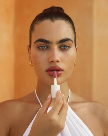 Close-up of a blue-eyed woman applying lip gloss with a wand. The background is a soft peach color, shot indoors with natural lighting.