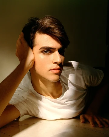 Close-up portrait of a young man wearing a white t-shirt, resting his head on his hand with dramatic lighting.