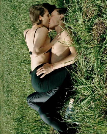 Two people embracing and kissing while lying amidst tall green grass in an outdoor meadow.
