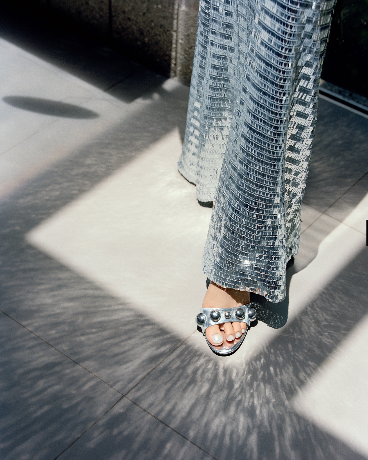 Woman's legs in silver sequin pants and studded heels standing on a gray tiled floor with geometric light and shadow patterns.