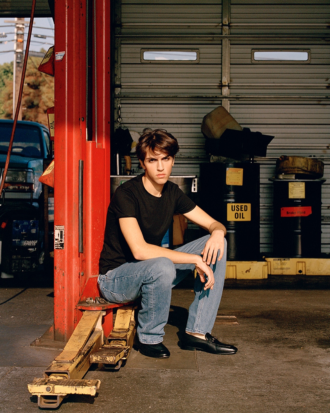 Adult male in black shirt and jeans sits on a red auto lift in an auto repair shop with a car and other equipment in the background.