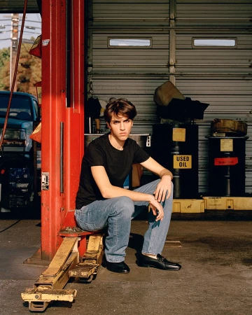 Adult male in black shirt and jeans sits on a red auto lift in an auto repair shop with a car and other equipment in the background.