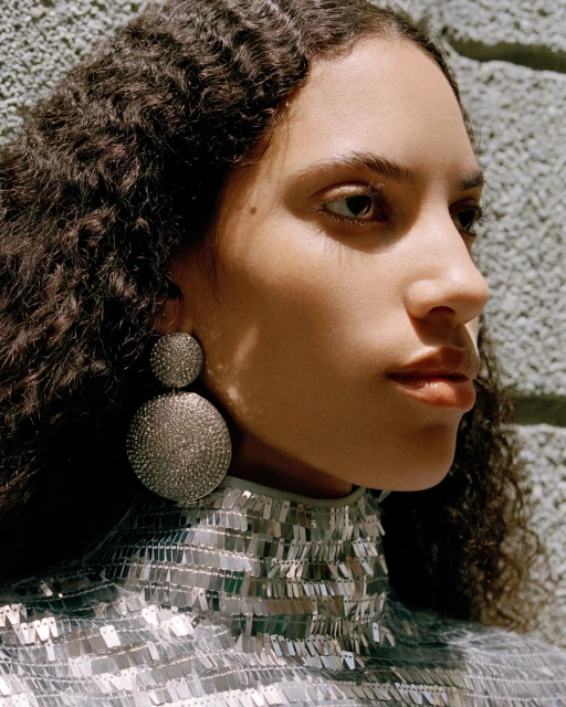 Close-up portrait of a woman with curly hair wearing large, round earrings and a silver, sequined top against a textured gray wall.