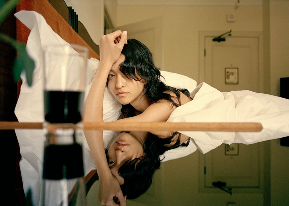 Portrait of a young adult female lying in bed under white sheets with her head on a pillow, reflected in a glass table.