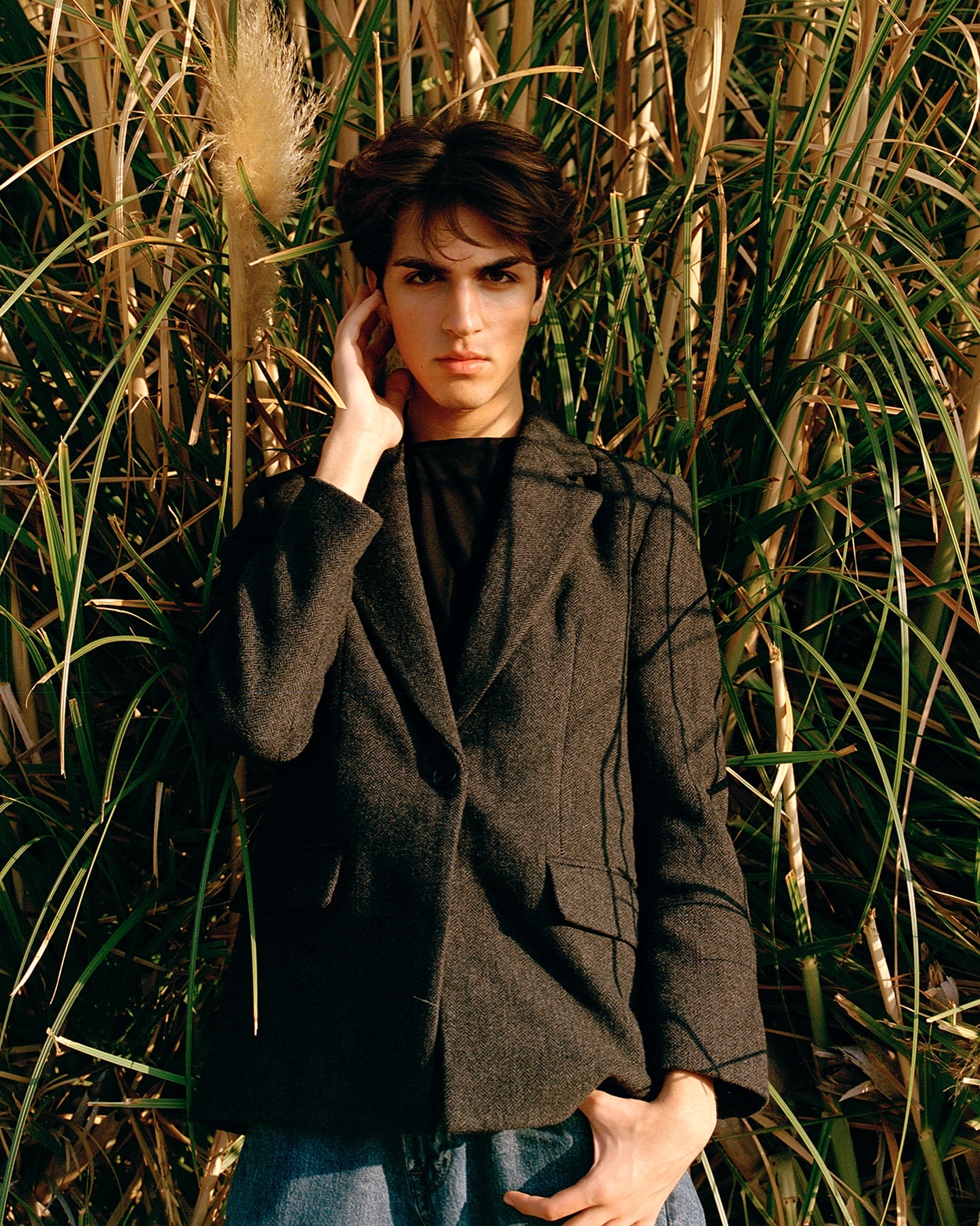 A young adult stands surrounded by tall grasses wearing a blazer, looking directly at the camera with a serious expression.