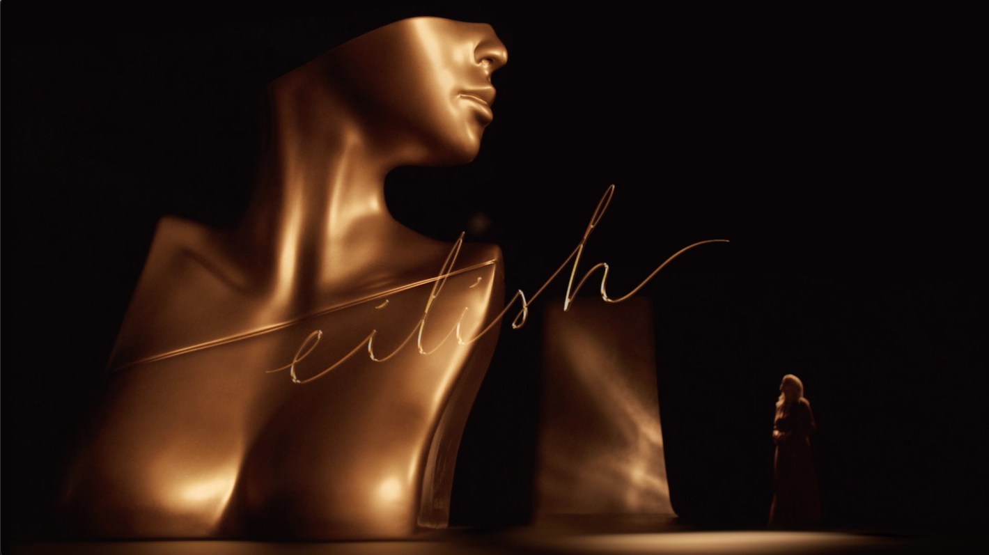 Golden bust sculpture with script 'fetish' in foreground, small female figure, soft lighting, dark background, high-contrast.