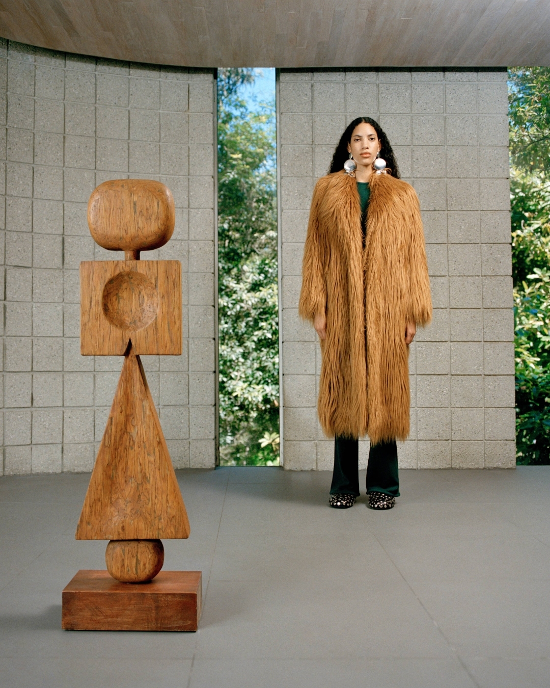 Woman in a fur coat standing next to a wooden sculpture in a concrete room with a window to greenery, fashion photography.