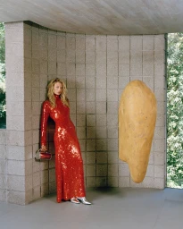 Woman in a shiny red sequin long-sleeved dress leaning against a grey concrete block wall holding a small handbag.