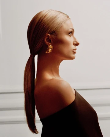 Profile portrait of a blonde woman wearing a dark off-the-shoulder top and large gold seashell earring against a white wall.