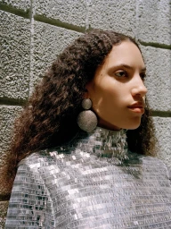 Portrait of a woman wearing a sparkling sequin top and large circular rhinestone earrings against a textured concrete block wall.