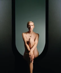 A blonde woman seated inside an arched window opening, wearing a delicate necklace against a muted background.