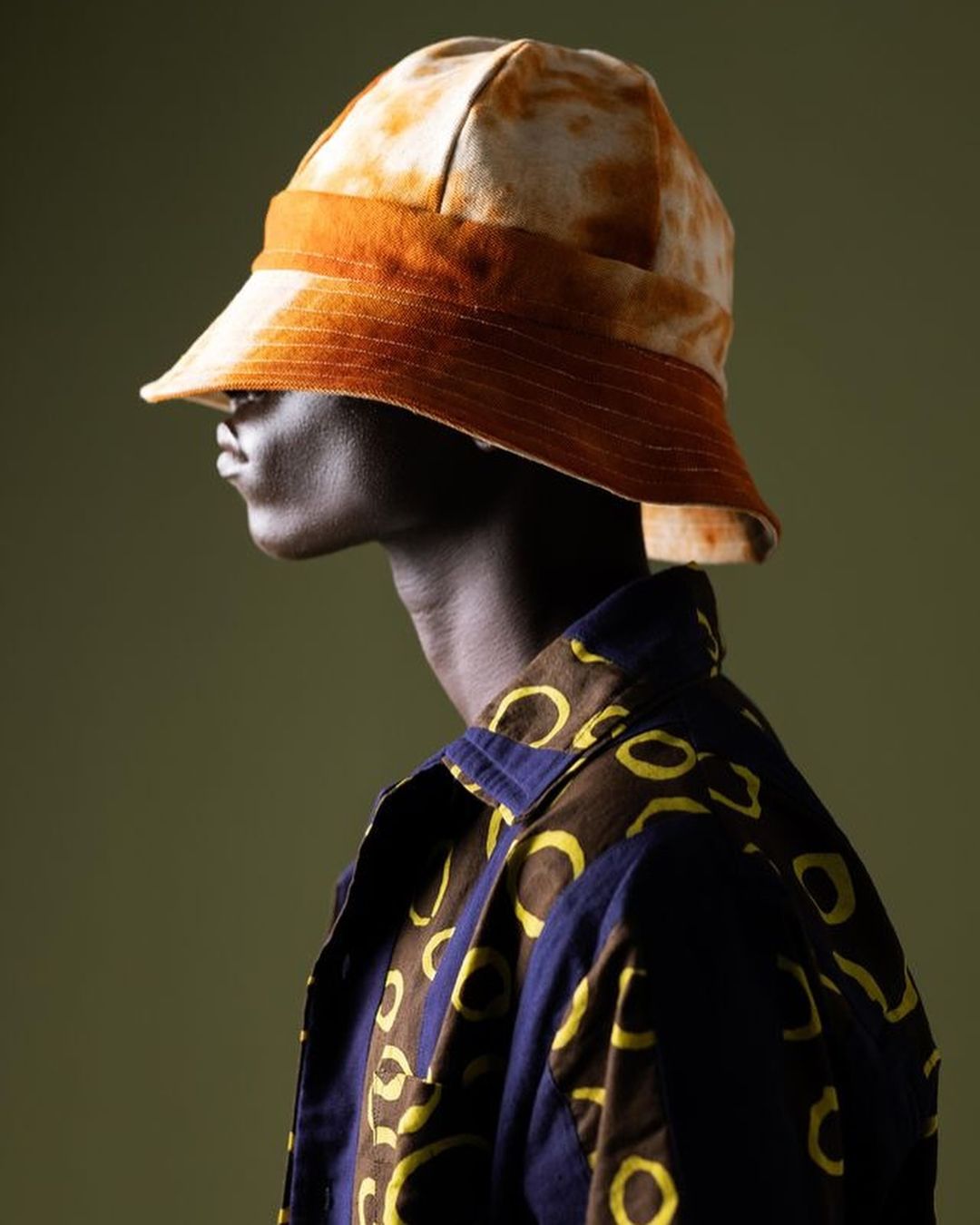 Profile shot of a dark-skinned adult male wearing a bucket hat with earth-tone designs and a patterned shirt against a green background.