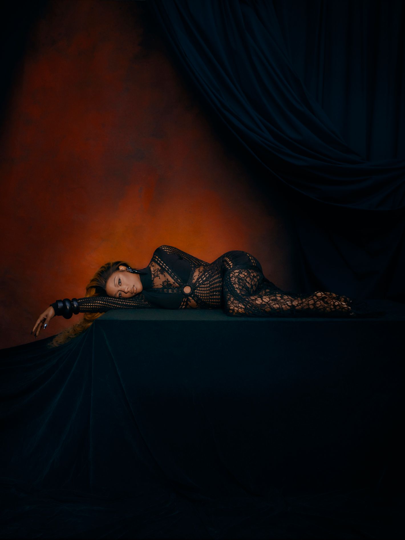 A woman in a lace catsuit is lying on a draped surface in a dimly lit studio with an orange and black background, in a fashion-style pose.